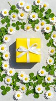 Yellow gift box with white ribbon surrounded by fresh daisies and clover leaves on neutral background for spring celebrations and special occasions.