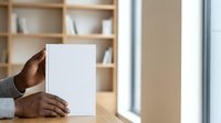 Young African American male professional holding blank white paper sheet at wooden desk with shelving background, suitable for business presentations and corporate communications.
