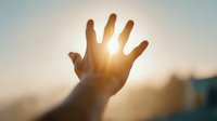 Hand reaching toward bright sunlight with warm golden rays shining through fingers against soft blurred background creating hopeful inspirational moment.