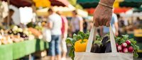 Man holding canvas tote bag filled with fresh vegetables at farmers market. Blurred background shows produce stands and shoppers browsing seasonal crops.