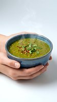 Hands holding ceramic bowl with green lentil soup garnished with herbs and chili flakes on white background for healthy cooking and nutrition concepts.