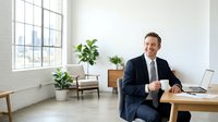 Confident male professional in dark business suit and tie sitting at wooden desk with laptop, holding coffee cup in modern minimalist office with large windows, potted plants, and natural light.