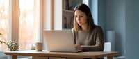 Young professional woman working on laptop at home office desk with natural sunlight streaming through windows for remote work and productivity.