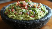 Fresh guacamole with diced tomatoes, onions and cilantro served in traditional dark stone molcajete bowl on wooden surface, authentic Mexican appetizer.