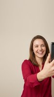 Happy young Caucasian woman in red shirt taking selfie with smartphone, smiling broadly while holding mobile device for social media content creation.