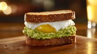 Toasted sandwich with mashed avocado, poached egg showing runny yolk on rustic wooden surface against warm bokeh background, appetizing breakfast composition.
