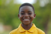 Young African American boy wearing yellow polo shirt smiling brightly at camera with natural green background, showing genuine happiness and confidence in casual outdoor portrait.