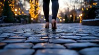 Female legs in athletic wear walking on cobblestone path at dusk, with bokeh lights and illuminated trees creating magical evening atmosphere in urban park setting.