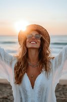 Young woman in straw hat and sunglasses enjoying sunset at beach, wearing white bohemian dress and layered necklaces, expressing joy and freedom.