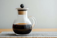 Glass coffee carafe with wooden lid and black handle on textured placemat, filled with freshly brewed dark coffee against neutral background.