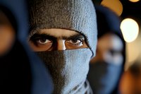 Close-up portrait of intense eyes visible through gray balaclava mask against dark background with blurred lights, conveying mystery and tension.