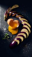 Sliced purple eggplant arranged in curved pattern with raw egg yolk and herbs on dark slate background for culinary cooking and recipe content.