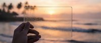 Hand holding transparent glass frame against tropical sunset beach with palm trees and ocean waves creating dreamy golden hour atmosphere for design concepts.