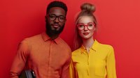 Diverse young professionals in bright orange and yellow shirts wearing stylish glasses pose against vibrant red background. Modern corporate diversity concept.