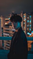 Young Asian male model in black jacket against urban night cityscape with bokeh lights, dramatic moody portrait with teal and orange color grading.