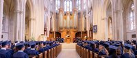 Graduation ceremony in gothic cathedral with students in caps and gowns seated in wooden pews facing ornate altar and pipe organ during commencement.