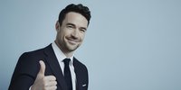 Confident businessman in navy suit giving thumbs up with friendly smile against light blue background, conveying success and approval.