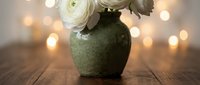 White ranunculus flowers in rustic green ceramic vase on wooden table with warm bokeh lights creating cozy home decor atmosphere for interior design.