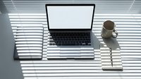 Laptop with blank white screen on desk beside coffee mug and notebook, sunlight stripes from blinds creating elegant shadow pattern.