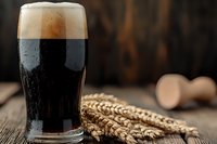 Dark craft beer in pint glass with creamy foam head alongside golden wheat ears on rustic wooden table, moody lighting creates dramatic beverage photography.