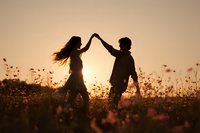Romantic couple silhouettes holding hands in wildflower meadow at golden sunset creating heart shape with arms for love and relationship concepts.