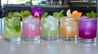Colorful tropical cocktails garnished with fresh mint leaves and edible flowers lined up on bar counter, refreshing summer drinks with crushed ice and citrus.