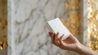 Hand holding blank white box against marble and gold textured background for product packaging mockup and branding presentation.