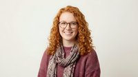 Young redhead woman with curly hair wearing glasses and burgundy sweater with scarf smiling at camera on neutral background for portrait photography.