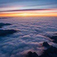 Dramatic sunrise above clouds with mountain peaks emerging through thick cloud layer creating ethereal landscape with golden and purple sky colors.