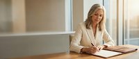 Professional blonde businesswoman in beige suit writing documents at modern office desk near window with natural lighting for corporate presentations.