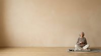 Middle-aged woman with gray hair, Caucasian ethnicity, sitting cross-legged on wooden floor in meditation pose wearing casual clothing. Minimalist interior with beige wall. Wellness and mindfulness concept.