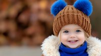 Cheerful blonde toddler wearing brown knit beanie with blue pom poms, blue scarf and cream winter coat smiles brightly against blurred autumn background.