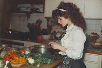Young woman with curly dark hair in vintage white blouse cooking in kitchen, preparing fresh vegetables and herbs on wooden cutting board, retro style home setting.