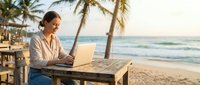 Young professional woman working on laptop at tropical beach cafe with palm trees during golden hour sunset creating peaceful remote work atmosphere.