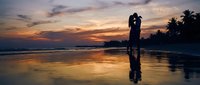 Romantic couple silhouette embracing on tropical beach at sunset with golden sky reflection on wet sand and palm trees creating intimate vacation moment.
