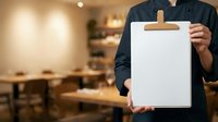 Person in dark business attire holding blank clipboard with wooden clip in modern restaurant or cafe setting, suitable for menu design, business proposals, hospitality concepts.