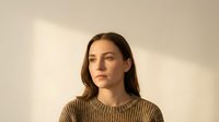 Young Caucasian woman in brown knit sweater with natural lighting and soft shadows against neutral beige wall background for lifestyle content.