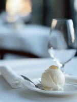Single scoop of vanilla ice cream served on white plate with elegant table setting, wine glass and rolled napkin in upscale restaurant atmosphere.