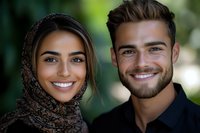 Happy young couple smiling outdoors with woman wearing traditional headscarf and man in casual attire against natural green background.