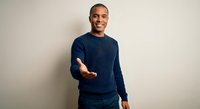 Smiling African American man in navy sweater extending hand for handshake against neutral background for business and professional concepts.