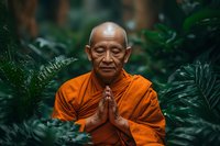 Senior Asian monk in orange robe meditating with prayer hands among lush green tropical plants, creating peaceful spiritual atmosphere for wellness and mindfulness practices.