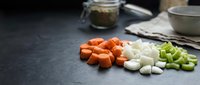 Fresh diced vegetables including orange carrots, white onions and green celery arranged on dark kitchen counter for cooking preparation and meal ingredients.