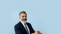 Middle-aged man with gray beard and dark hair, South Asian ethnicity, wearing navy blazer and white shirt against light blue background, professional business portrait.