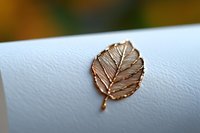 Delicate rose gold leaf brooch pin on white textured paper background, showing intricate natural vein pattern detail in metallic finish. Macro jewelry photography.