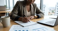 Professional African American businesswoman writing notes in modern office workspace with laptop and financial charts on desk.