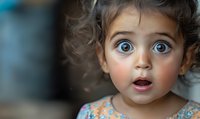 Young child with striking blue eyes and curly hair expressing amazement and wonder, mouth open in surprise against blurred background. Emotional portrait capturing innocence.