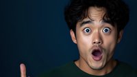 Young Asian man with shocked surprised expression and wide open mouth pointing finger against dark background. Dramatic studio lighting creates emotional portrait.