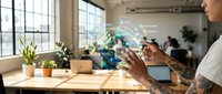 Professional woman with tattoo interacting with holographic data visualization in modern office workspace with natural lighting and collaborative environment.