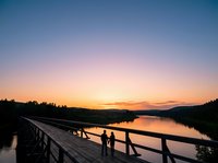 Silhouettes of two people standing on wooden bridge railing at sunset, overlooking calm river with forested mountains in background, golden and pink sky reflecting on water during dusk.