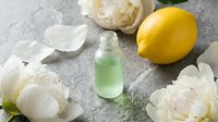 Bottle of green liquid surrounded by fresh lemons and white peony flowers on gray marble surface for cosmetics, skincare, or wellness product photography.